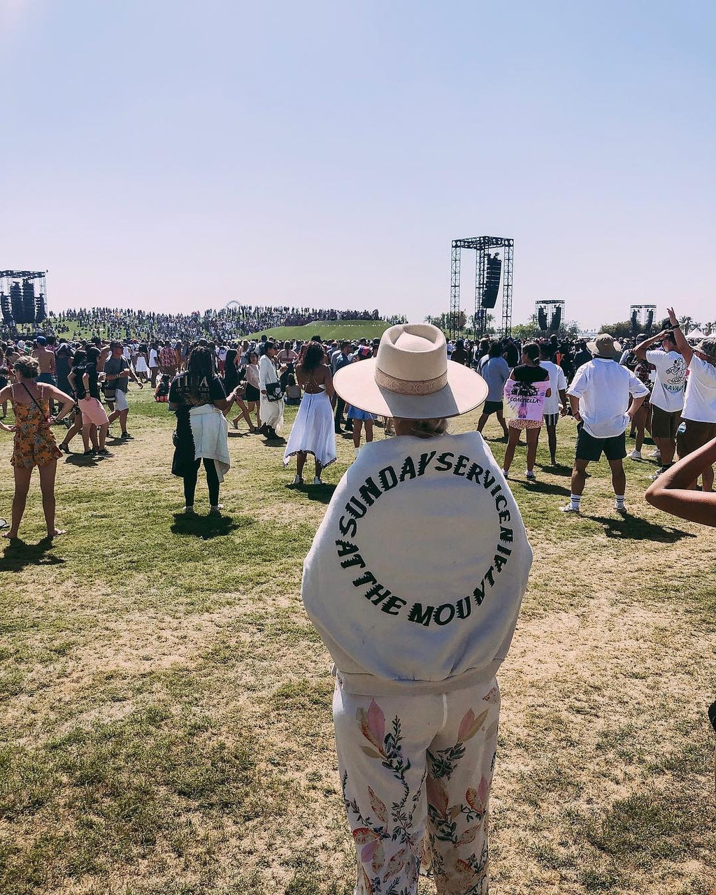 Person in a wide-brim hat wearing a white hoodie at a crowded outdoor event with a stage.